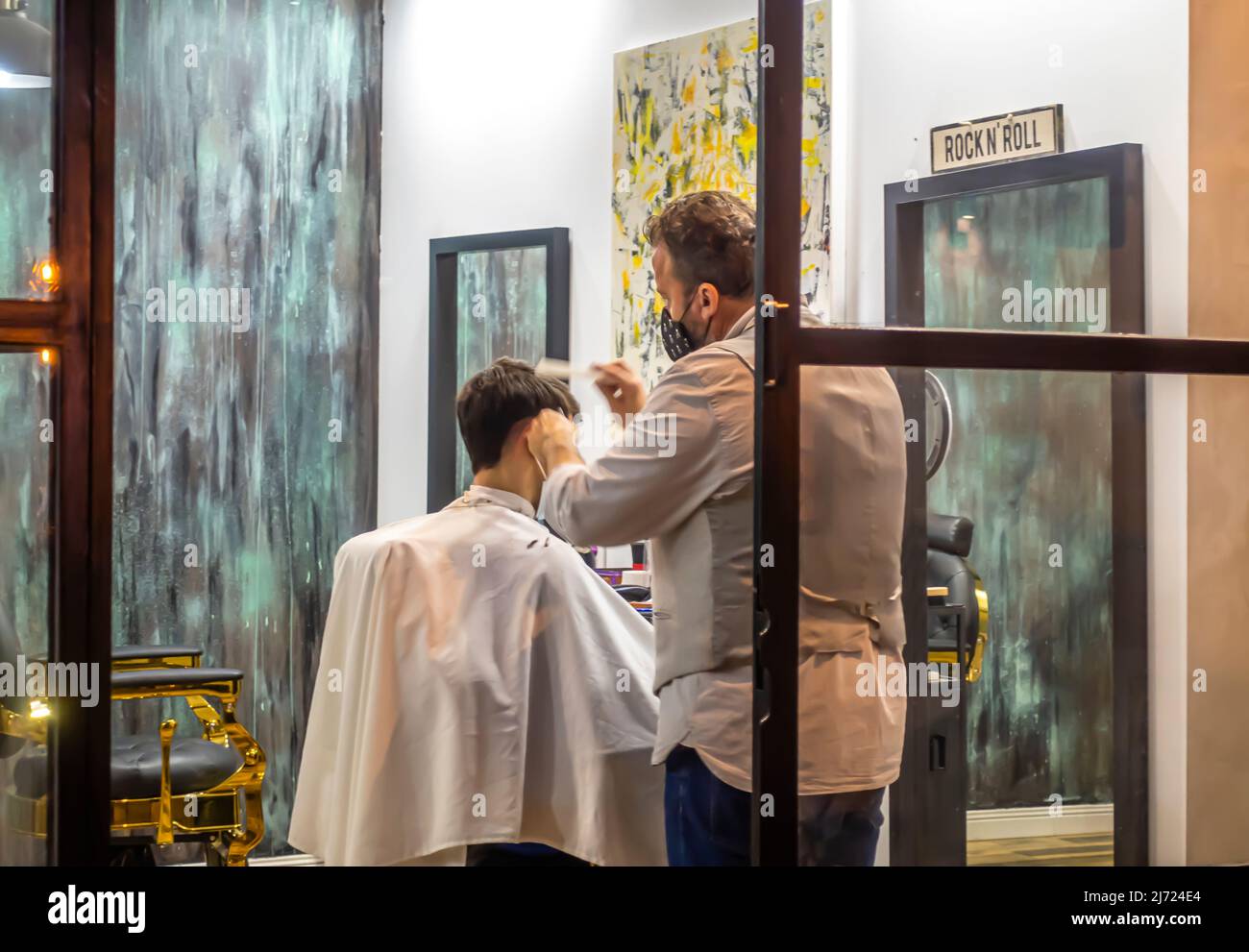 A barber in face mask working on a haircut in male barbershop in Seville, Andalucia, Spain. Sevillan Spanish hairdressers doing haircut hairdo. Stock Photo