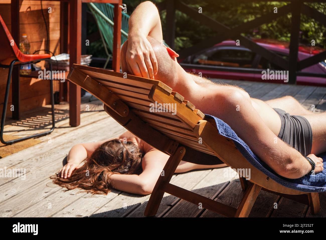 Young man resting on wooden terrace, view from behind - anonymous woman sunbathing over floor in background Stock Photo