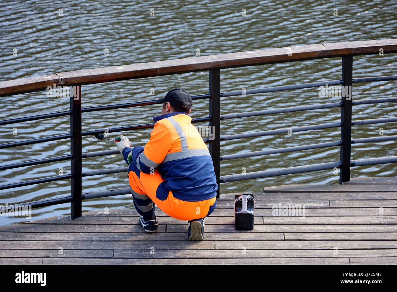 Worker in uniform paints the metal railing on a lake or river embankment. Repair works in spring park, preparing for the beach season Stock Photo