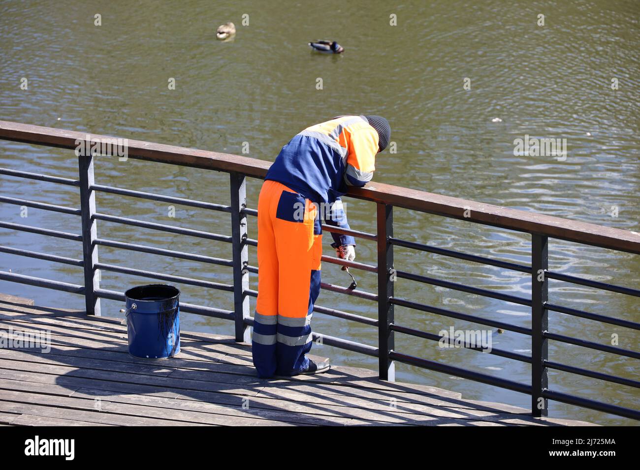 Worker in uniform paints the metal railing on a lake or river embankment. Repair works in spring park, preparing for the beach season Stock Photo