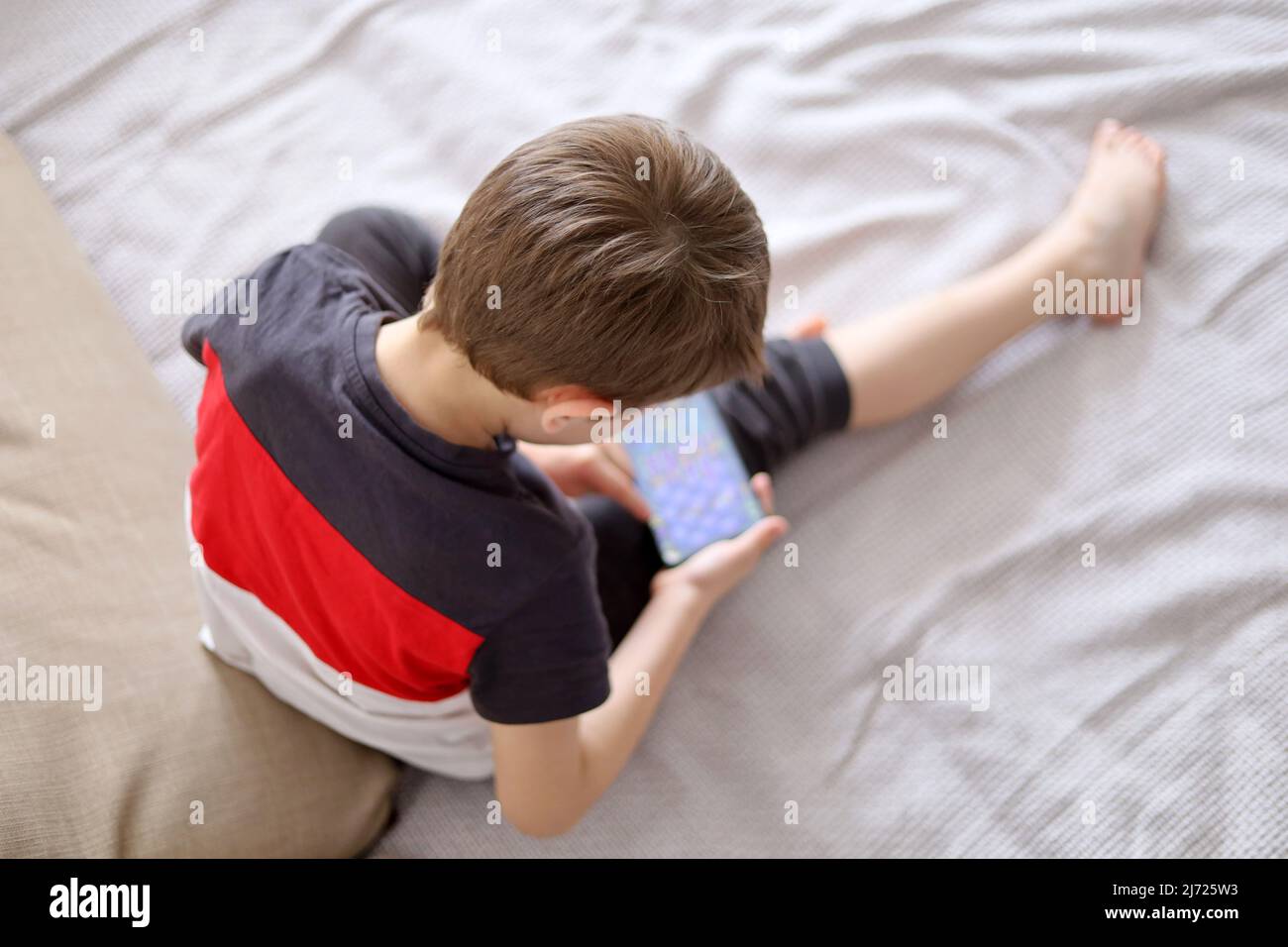 Little boy playing mobile game on smartphone sitting on a sofa, top view. Child leisure at home, video gaming addiction Stock Photo