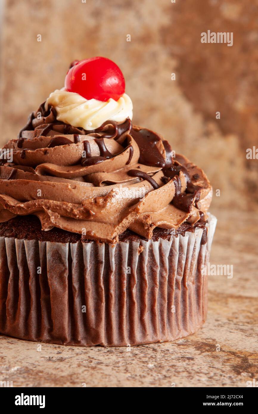 Still life closeup of a delicious chocolate cupcake with swirls of frosting and drizzled chocolate sauce, topped with a maraschino cherry. Stock Photo