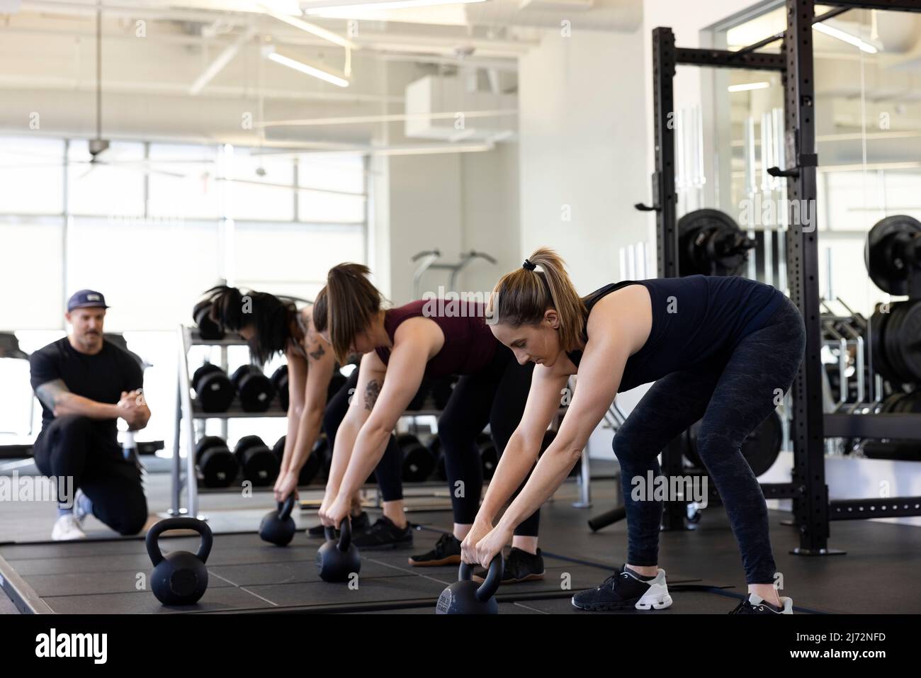 Three woman in a row bent over doing kettlebell exercise while personal trainer looks on in background. Stock Photo