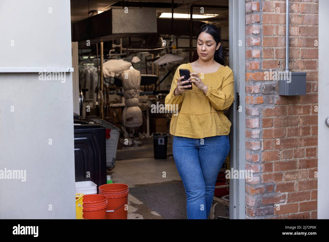 Young woman working in dry cleaners taking break in back room and using cell phone. Stock Photo