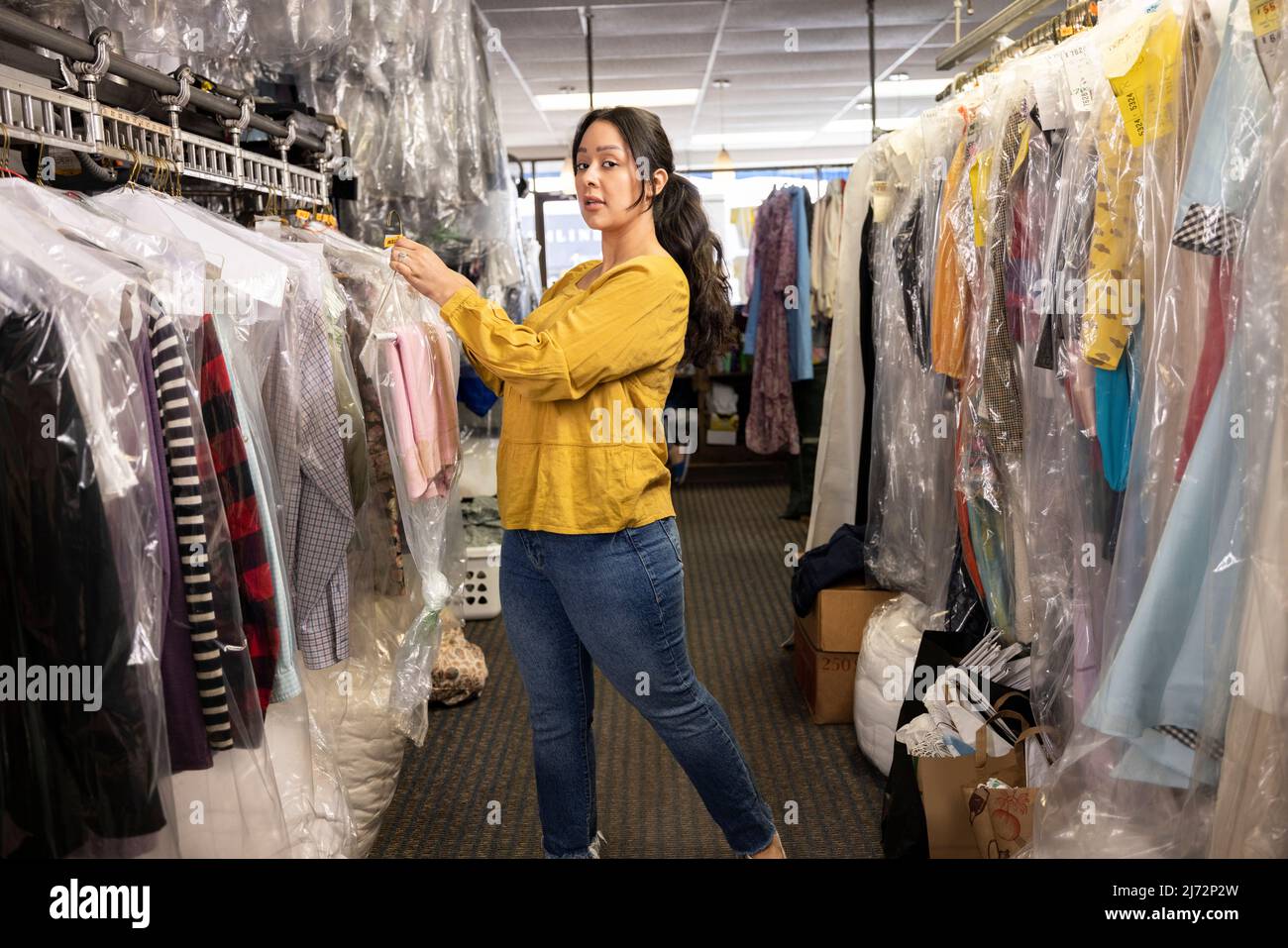 Side view of mid adult woman looking at camera and putting clothes away on clothing rack Stock Photo