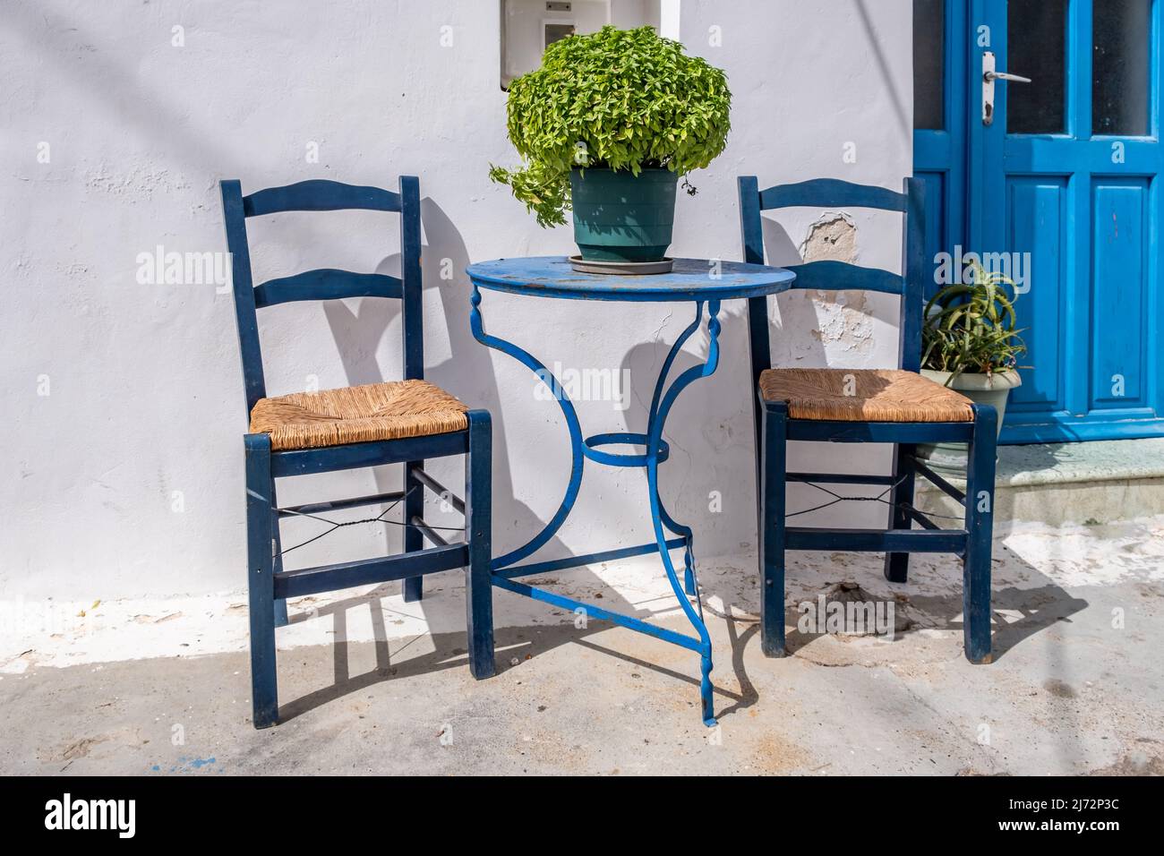 Empty blue round metal table and two wooden chairs, white traditional building wall. Outdoor cafe in a Greek island Chora, Greece. Summer sunny day. Stock Photo