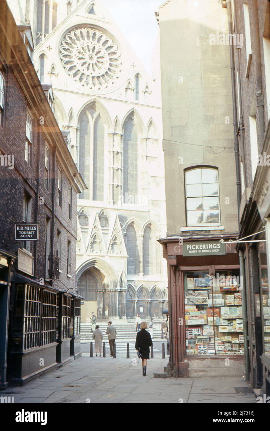 The southern façade of York Minster from Minster Gates, York, North Yorkshire.  1967. Shops are visible in the foreground including ‘R.B. Mills Tourist Agent’ and ‘Pickerings Hobby & Model Shop’. The latter has an interesting range of model kits displayed in its window. Stock Photo