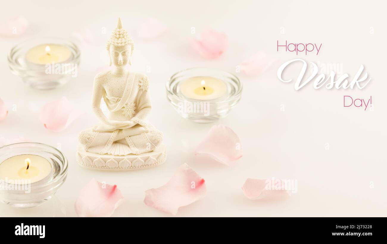 Vesak, Buddha Day holiday card. Buddha statue, blurry rose petals and candles on a white background with inscription Happy Vesak Day. Soft image and s Stock Photo