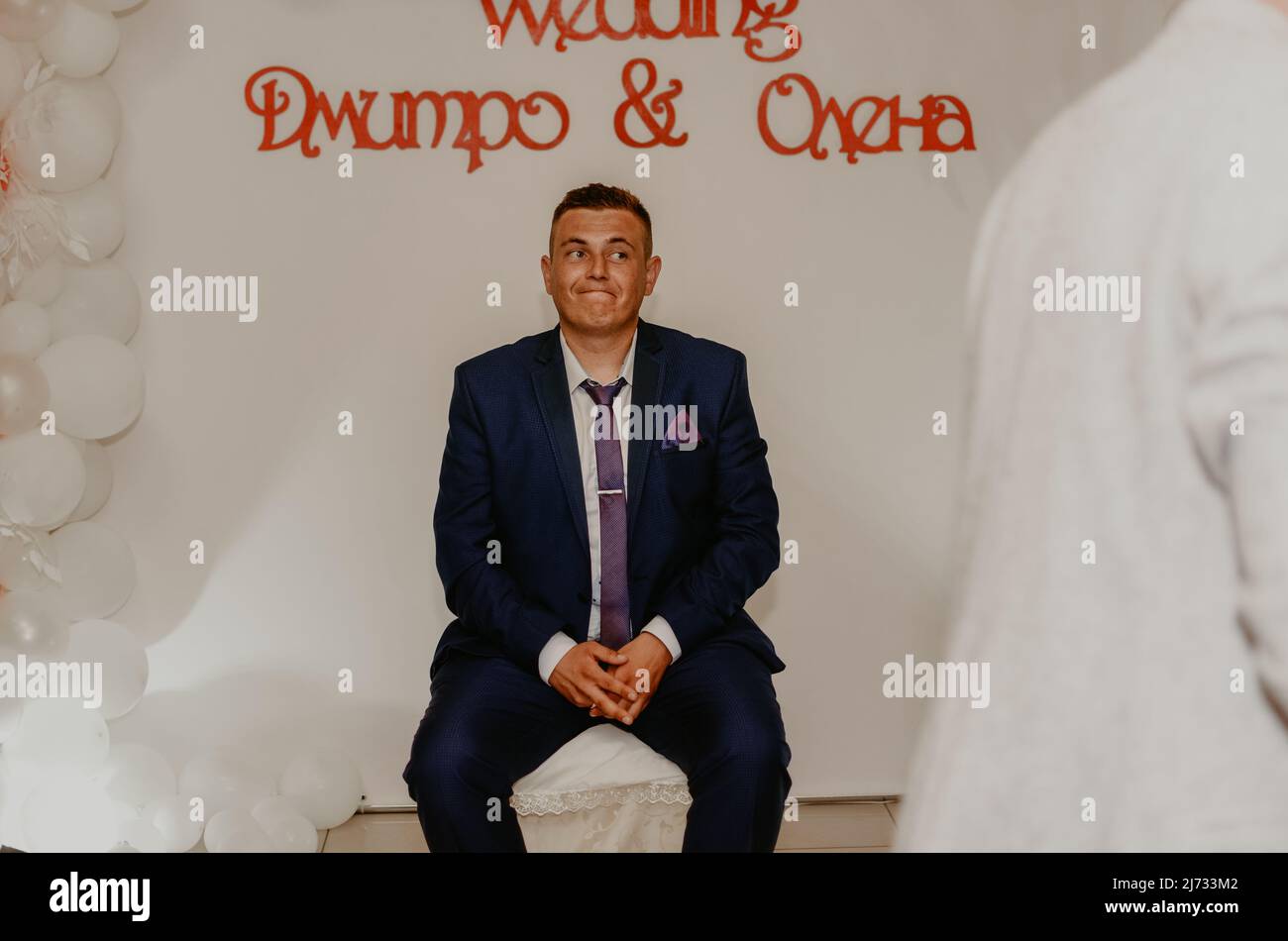 a man in a suit sitting on a chair puzzled. the groom the husband sits and waits for the bride. young man experiences strange emotions on face. Slavic Stock Photo