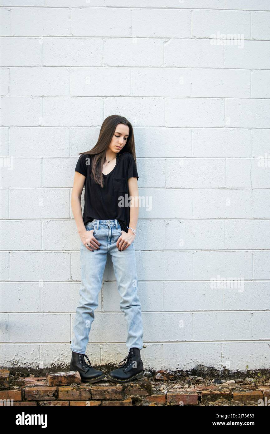 A teenage girl with long hair wearing black, denim and combat boots leaning against an old cinder block wall and looking down with attitude Stock Photo