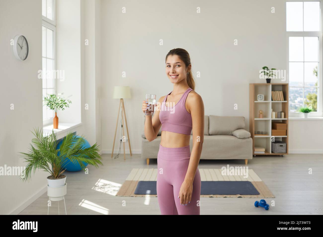 Lifestyle portrait of slender young fitness woman holding glass of water standing in living room. Stock Photo