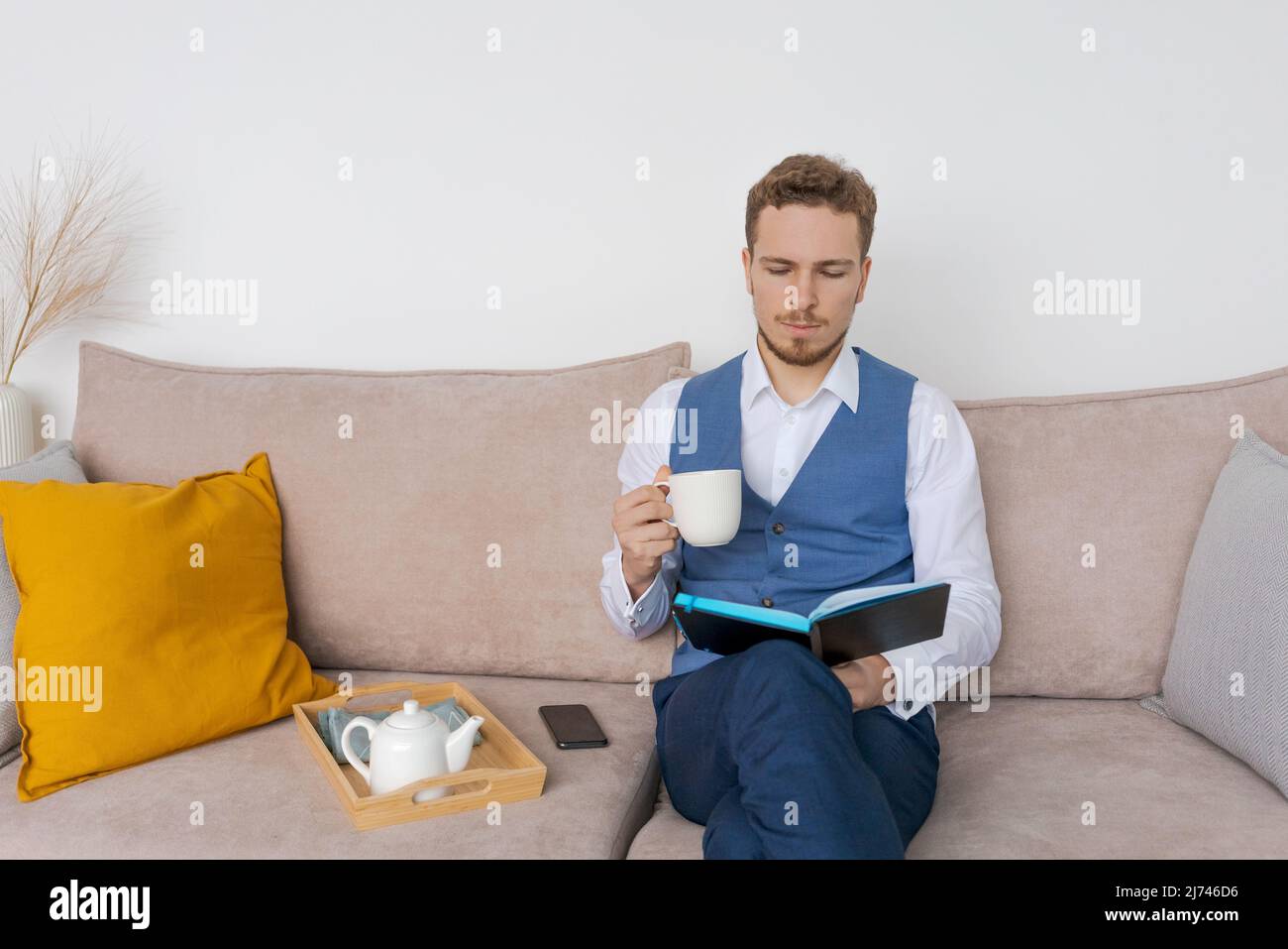 Pensive businessman sits on sofa with diary in his hand, thinks about plans in a blue bearded guy suit. Plans his daily routine and business meetings. Record keeping and planning concept Stock Photo