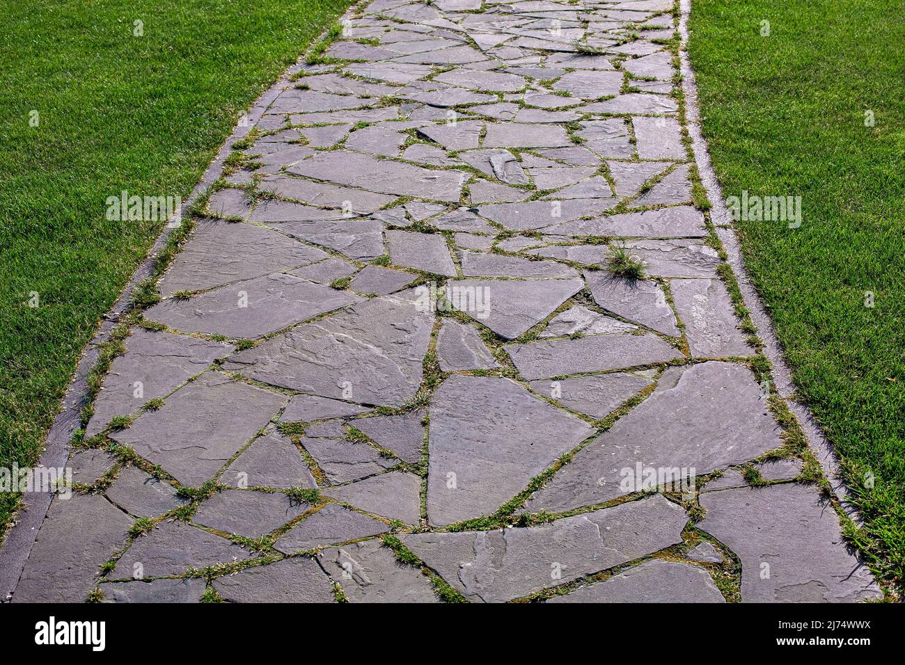 curved garden path made of natural stone paved with different size rough rock overgrown with grass in a park with a green lawn close-up of the turn wa Stock Photo