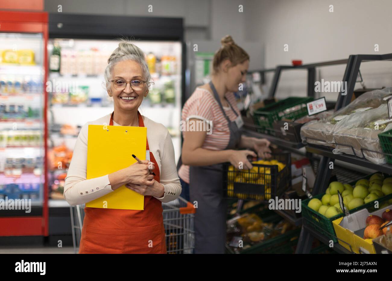 Confident senior shop assistant in supermarket in vegetable shell, in bakcground is her colleague filling stock. Stock Photo