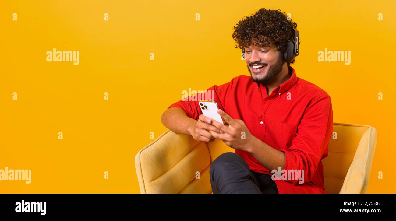 Studio portrait of young middle eastern guy in wireless headphones isolated on orange wall, looking at mobile phone, enjoying listening to favorite music soundtrack Stock Photo