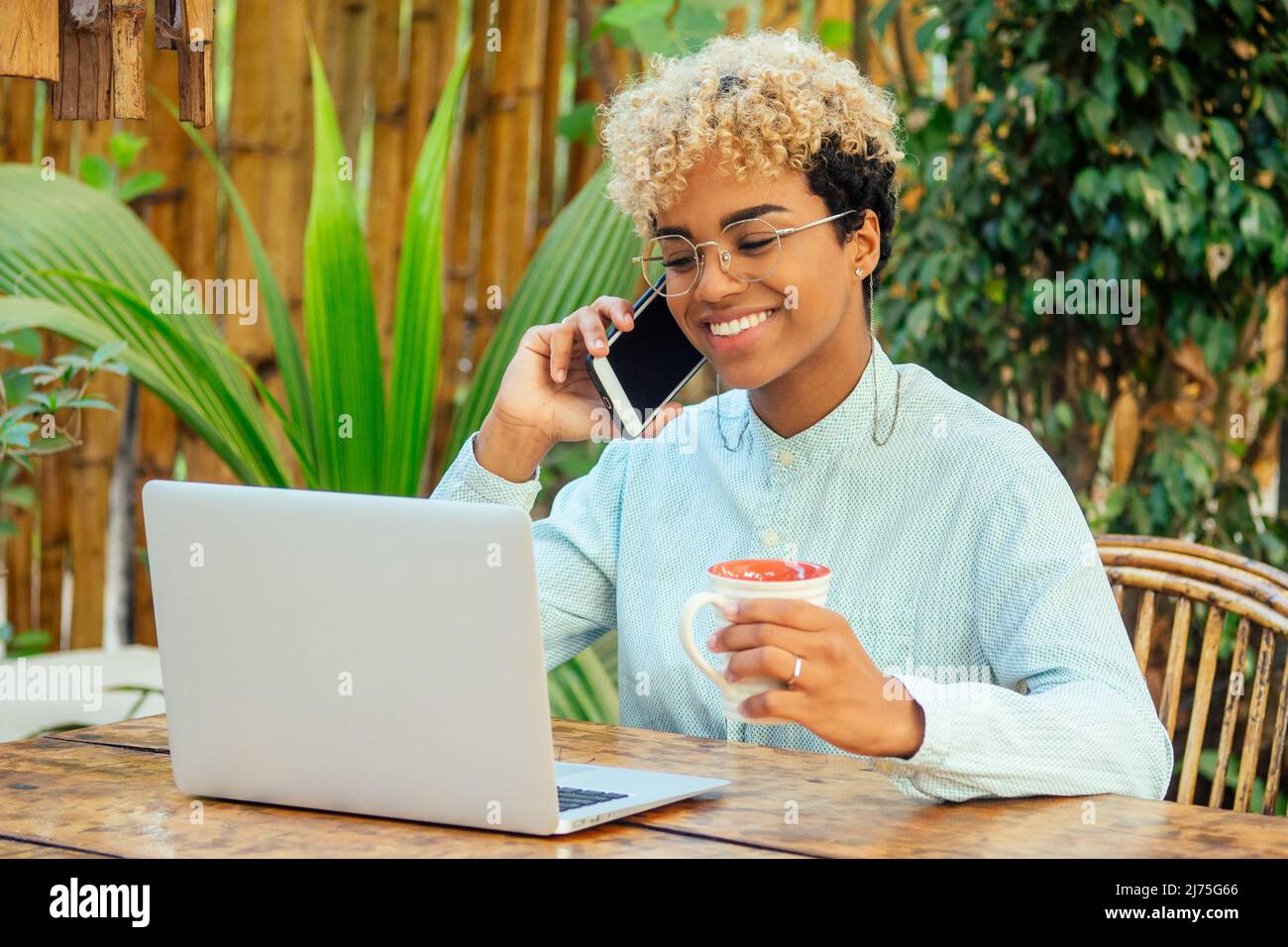 latin mixed race curly woman looking at laptop and smiling at tropical beach cafe with tea cup. She buying airline tickets online Stock Photo