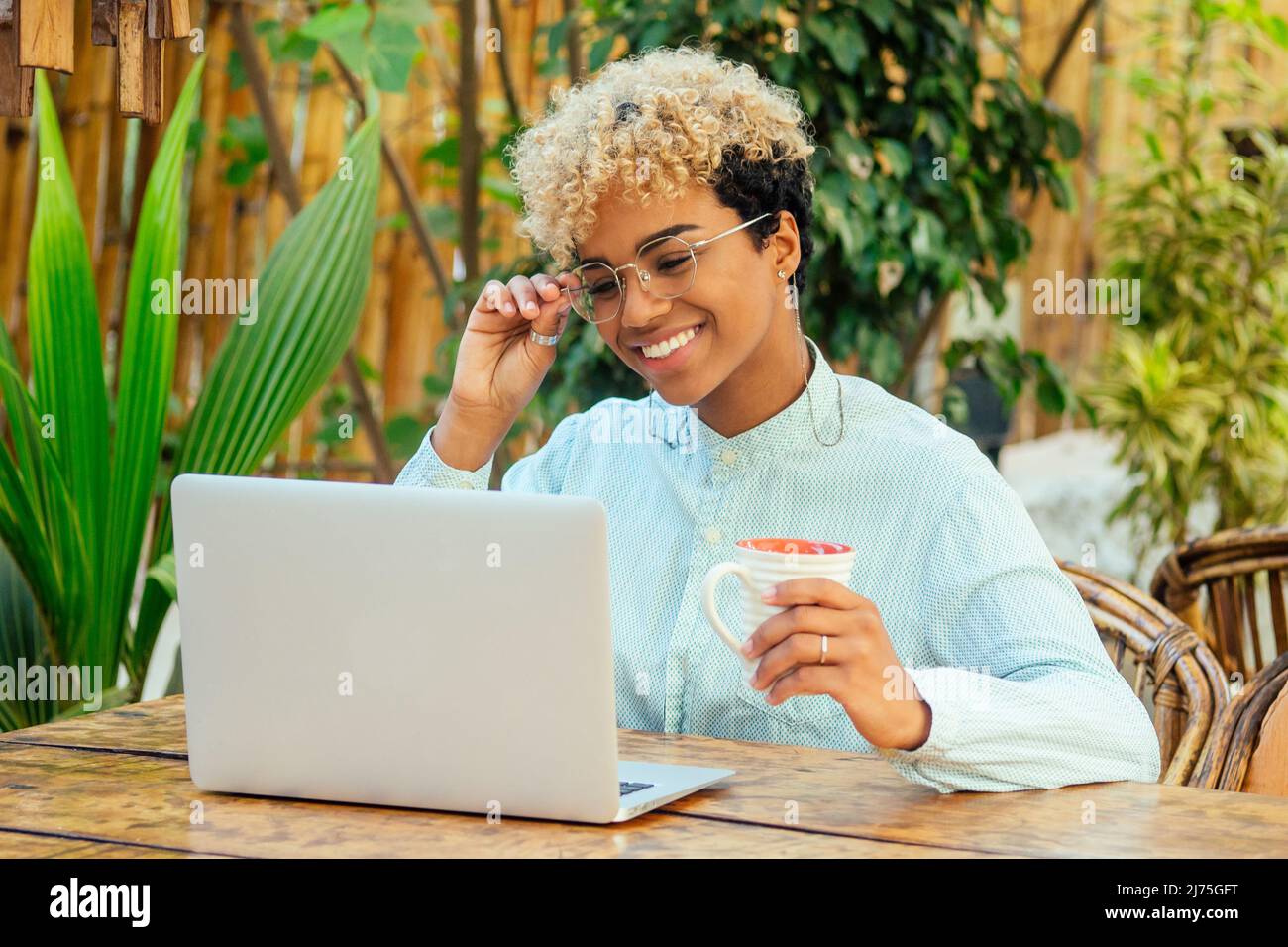 latin mixed race curly woman looking at laptop and smiling at tropical beach cafe with tea cup. She buying airline tickets online Stock Photo