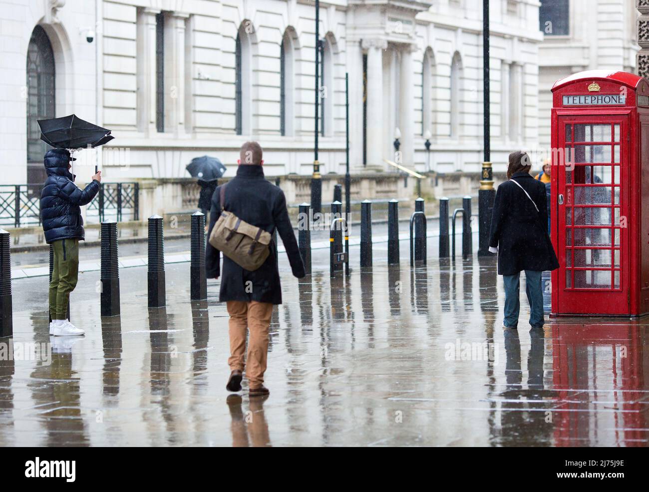 A person whose umbrella is seen blown by the wind takes a photograph of his company as a pedestrian walks past them in Westminster, central London. Stock Photo