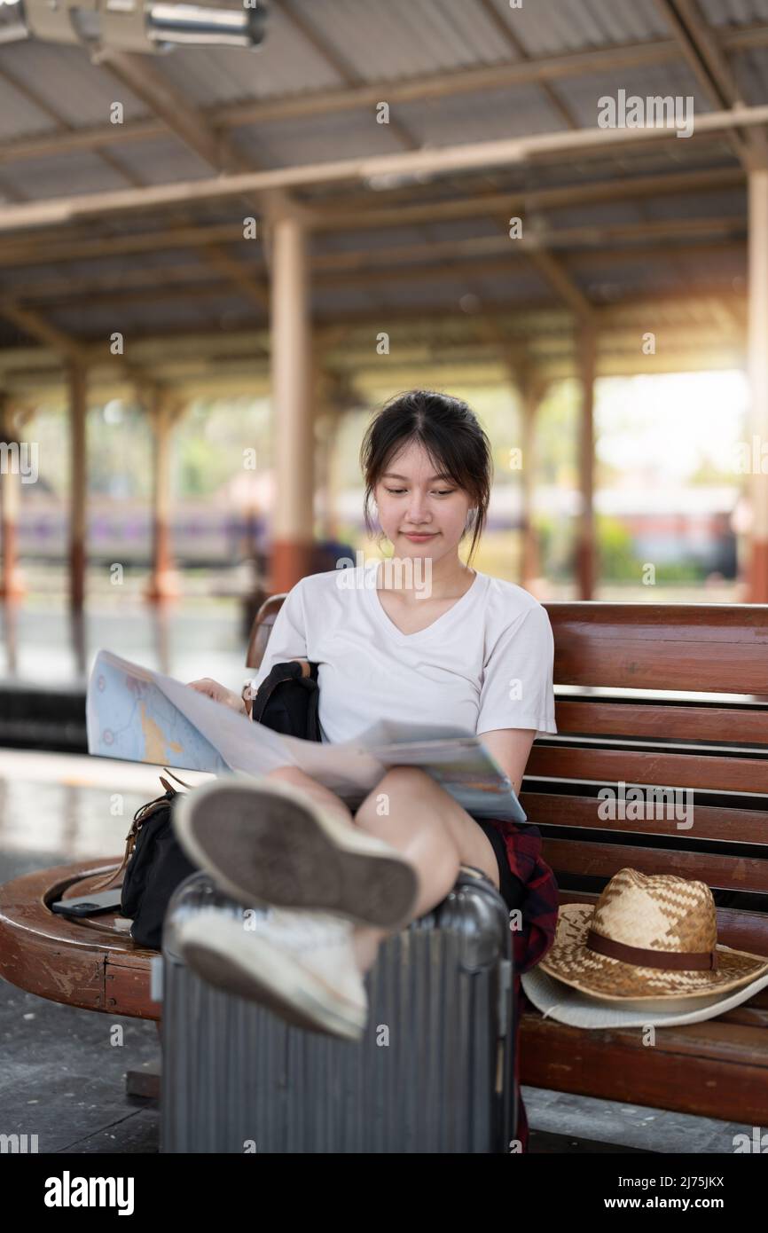 Happy young asian woman traveler or backpacker using map choose where to travel with luggage at train station, summer vacation travel concept Stock Photo
