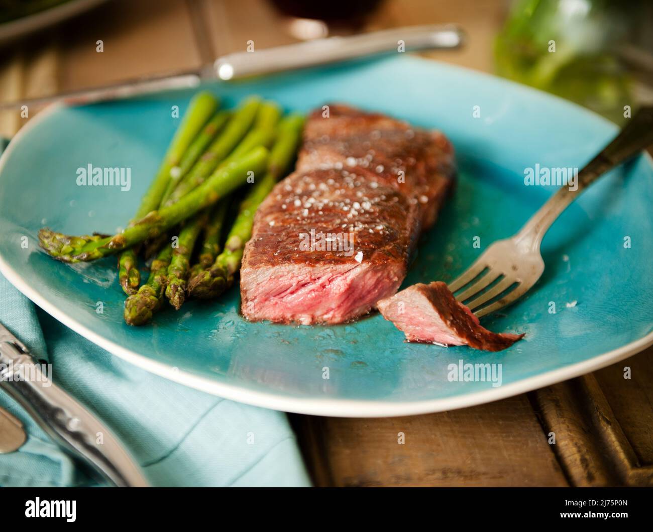 Strip Steak with a Piece Pierced on a Fork; Served with Asparagus on a Blue Plate Stock Photo