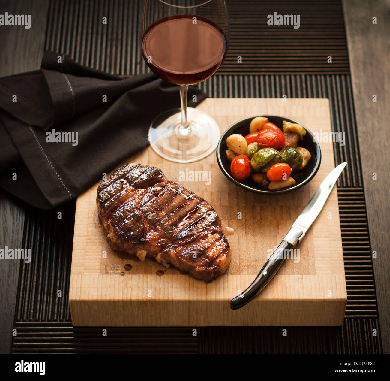 Whole Steak on a Cutting Board with a Bowl of Roasted Vegetables and a Glass of Red Wine Stock Photo