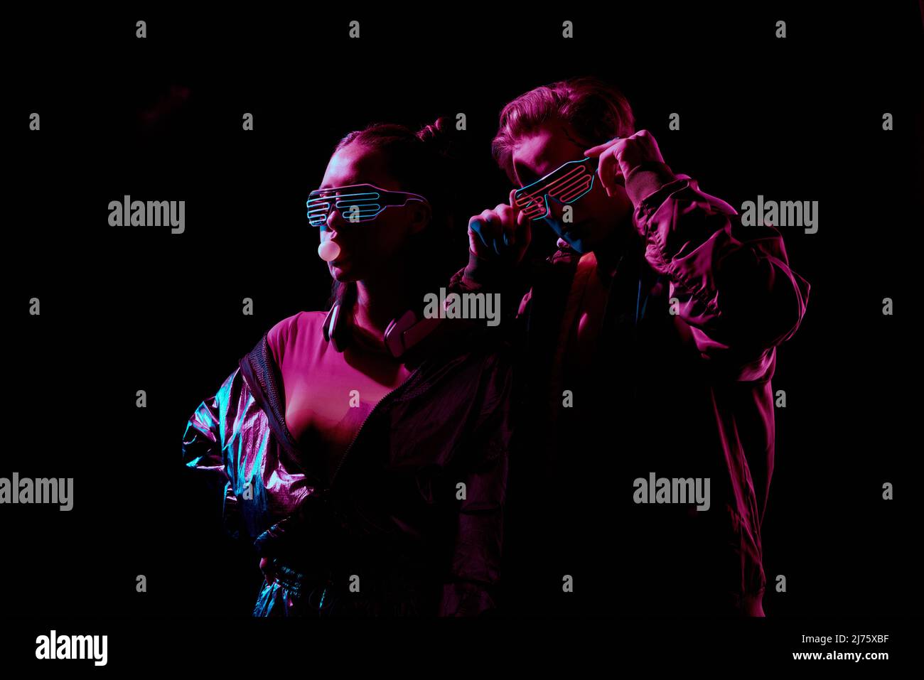Cool young couple in flashing LED goggles chewing gum and posing in darkness under red light Stock Photo