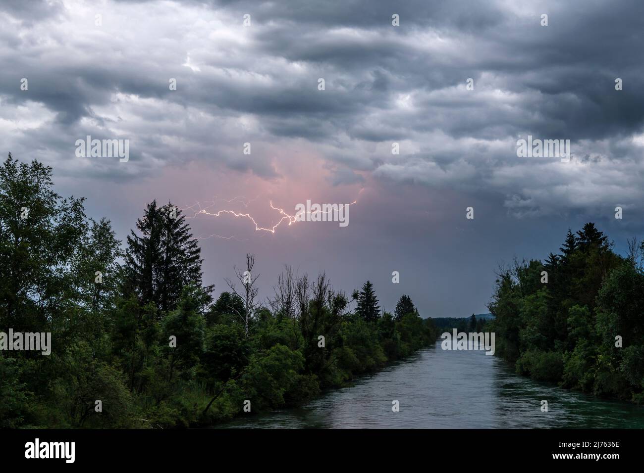 Lightning flashes in the sky above the Loisach, a well-known Bavarian river near Kochel am See, among other places. Thick clouds and the wildly overgrown course of the river contribute to the gloomy atmosphere. Stock Photo