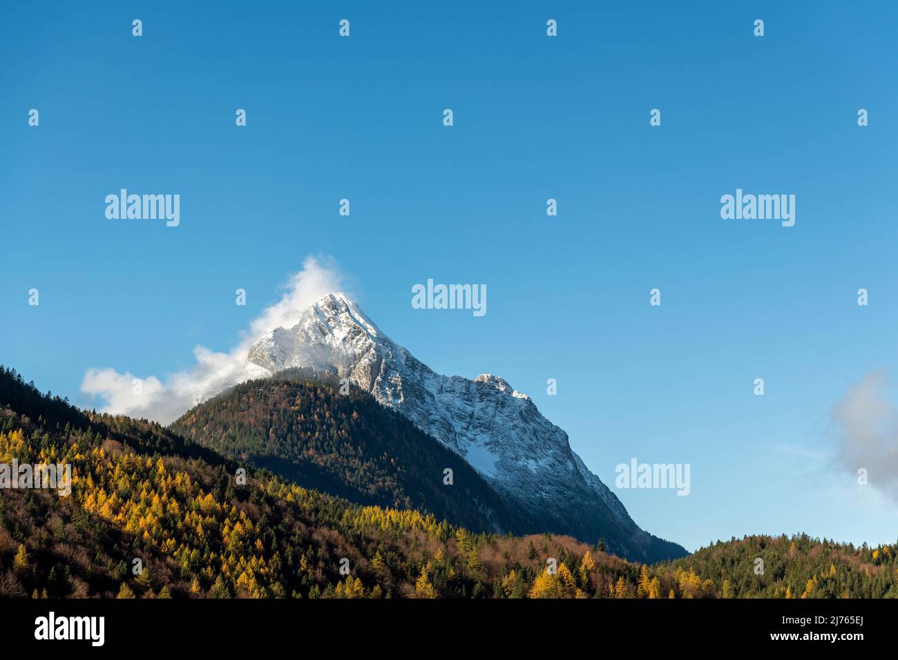 The Wetterstein, part of the mountain range of the same name between Garmisch-Partenkirchen and Mittenwald, seen from the village in autumn leaves and blue sky, with snow on the mountain. Stock Photo