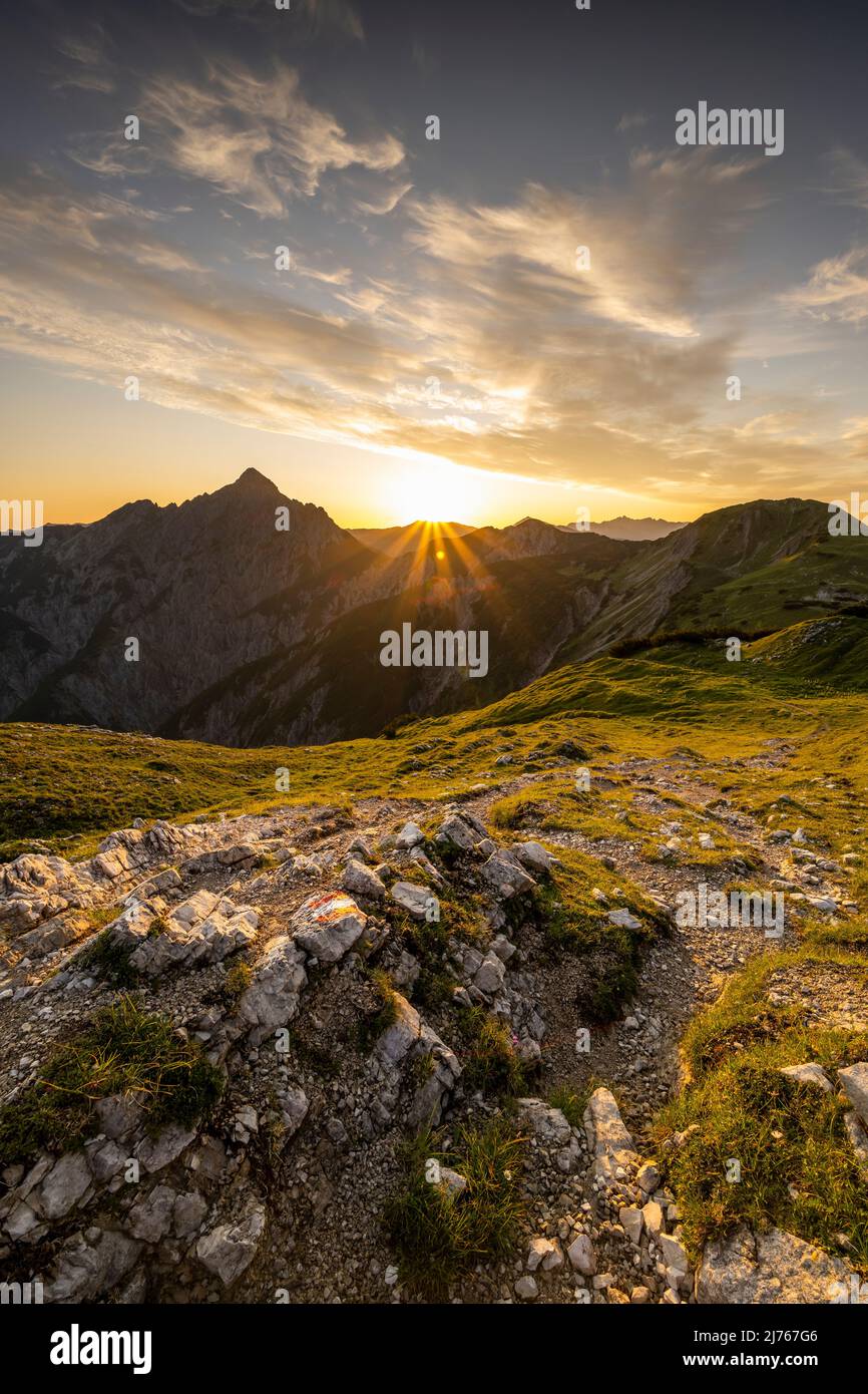 Hiking trail with trail markings, in the background the Mondscheinspitze with a great sun star with light colored clouds in the otherwise blue sky and golden, green mountain meadows. The trail leads over a ridge towards Plumsjochhütte from Kompar in the heart of the Karwendel. Stock Photo