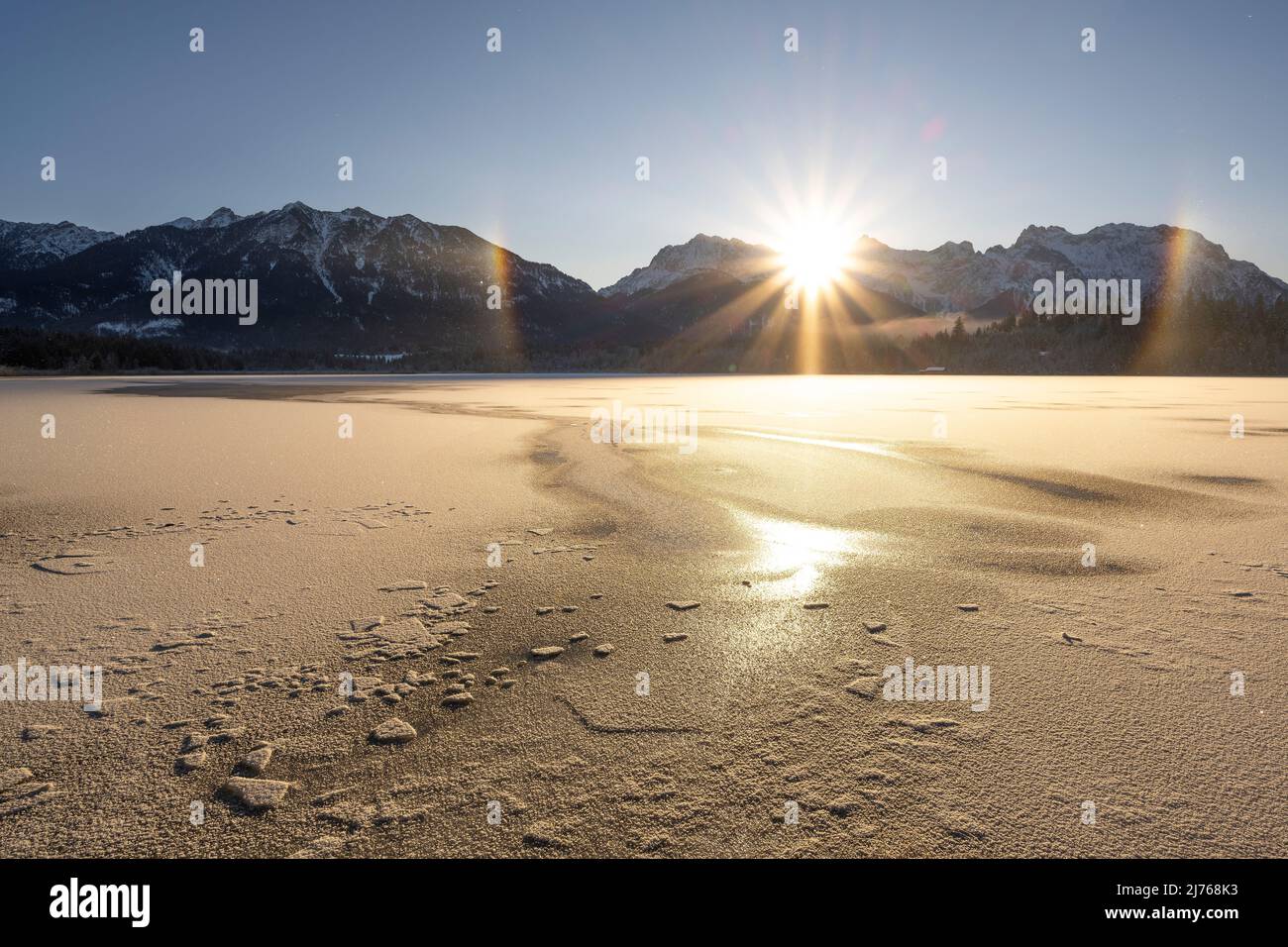 During sunrise, a rainbow formed by small ice crystals appeared against the light of the sun on the shore of the Barmsee. A small moor lake in the German Alps. In the background the Karwendel, in the foreground tracks in the ice. Stock Photo