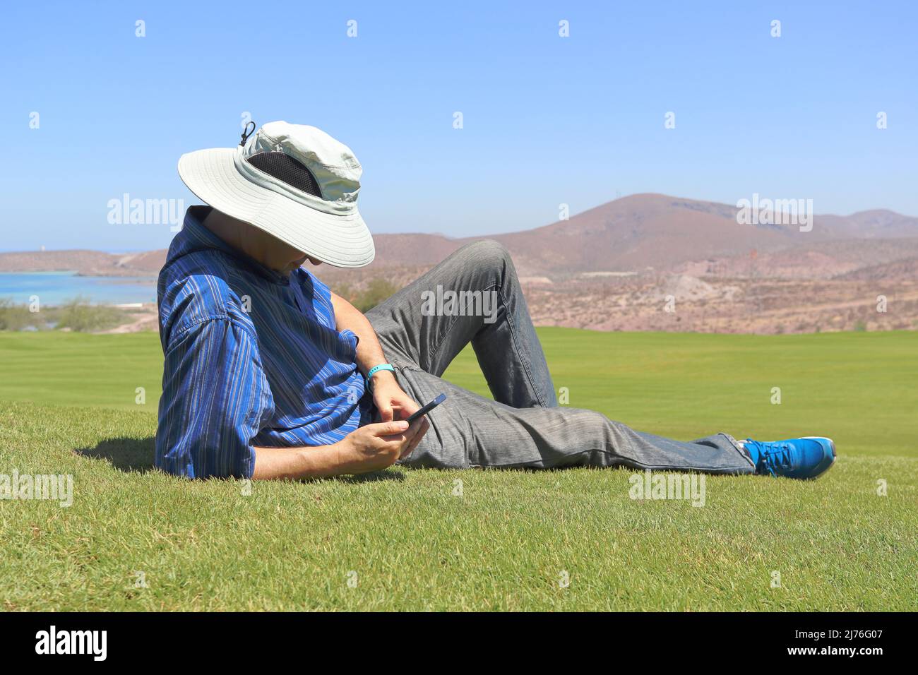 A man lying in the grass, relaxes while looking at his mobile phone. Stock Photo