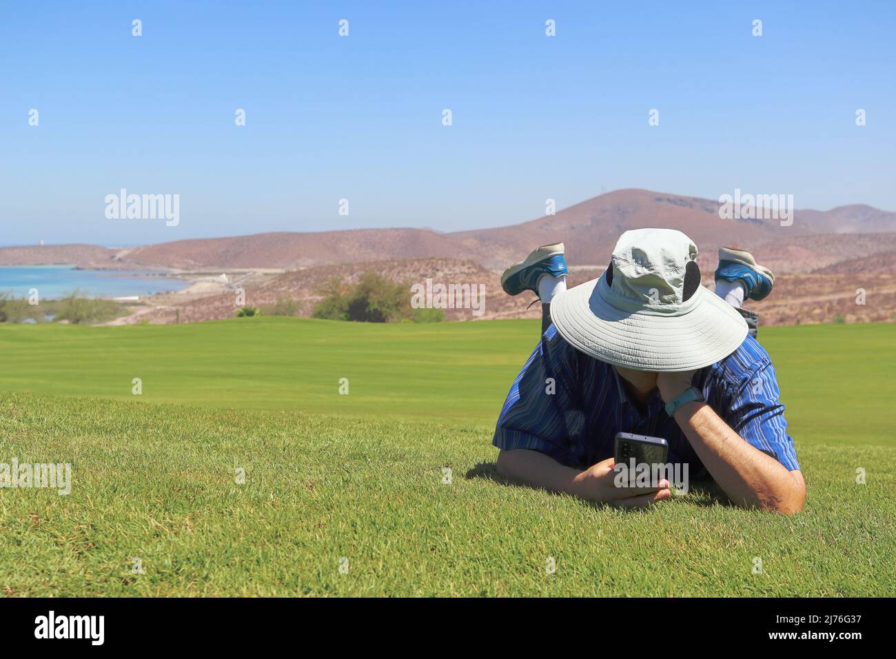A man lying in the grass, relaxes while looking at his mobile phone. Stock Photo