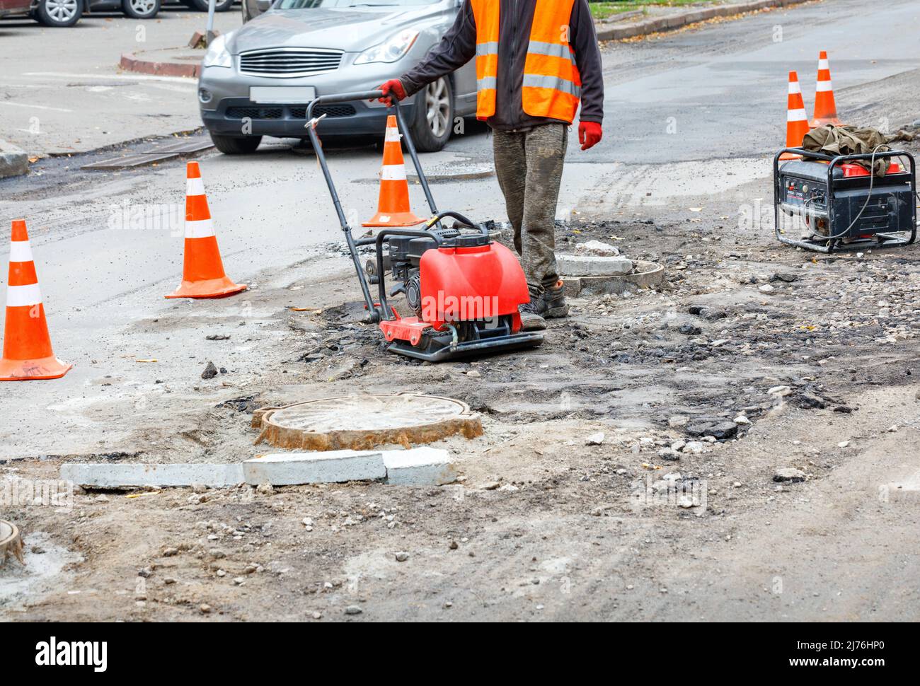 A road service worker rams a section of the carriageway fenced with orange traffic cones near sewer manholes with a gasoline rammer. Stock Photo