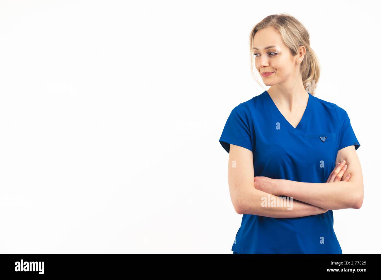 European blonde female helath care worker in a navy blue medical uniform standing with her arms crossed on her chest looking away. Studio shot on white background, copy space. High quality photo Stock Photo