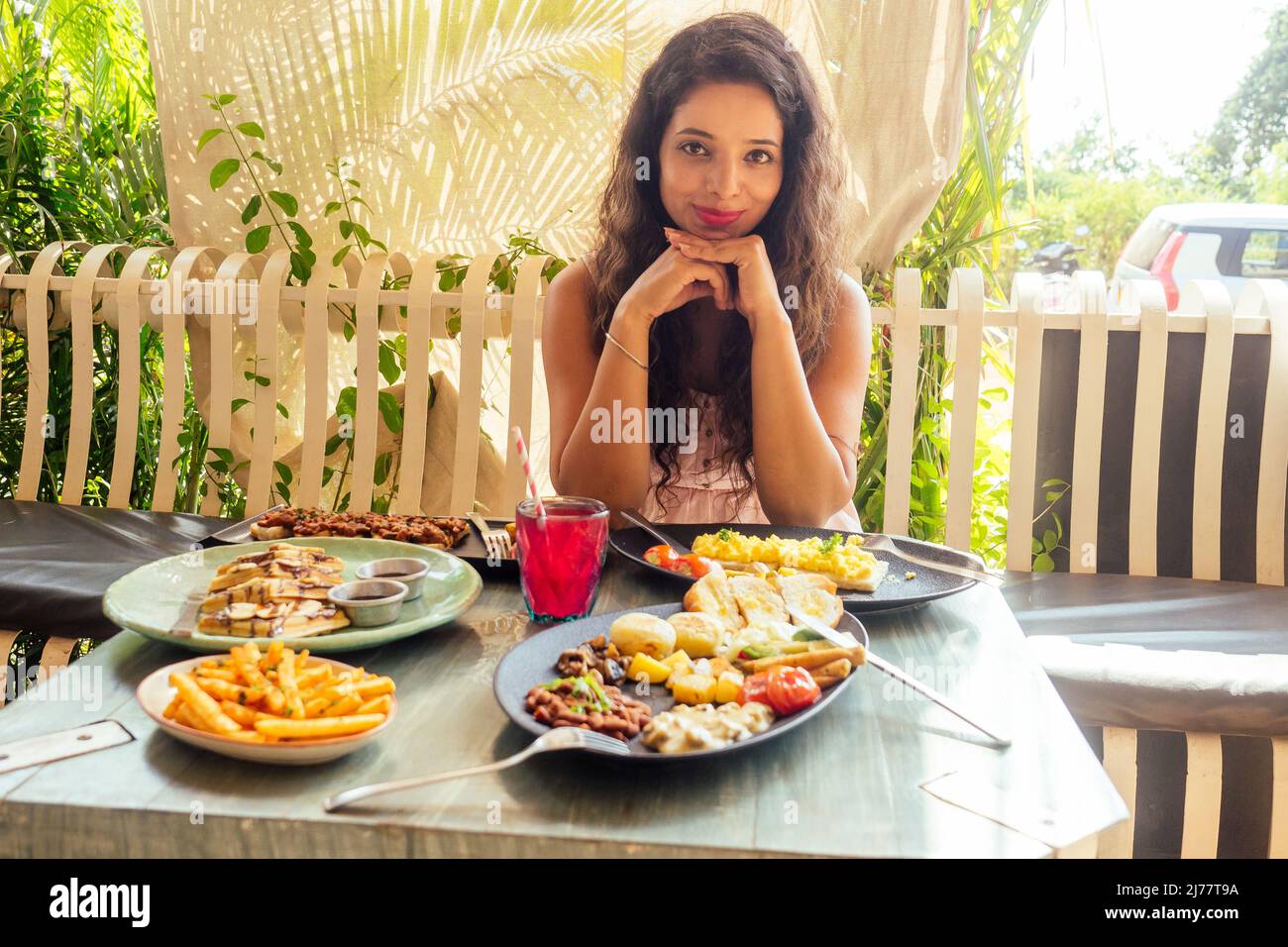indian pretty woman in pink dress and curly hairstyle sitting in summer tropical cafe Stock Photo