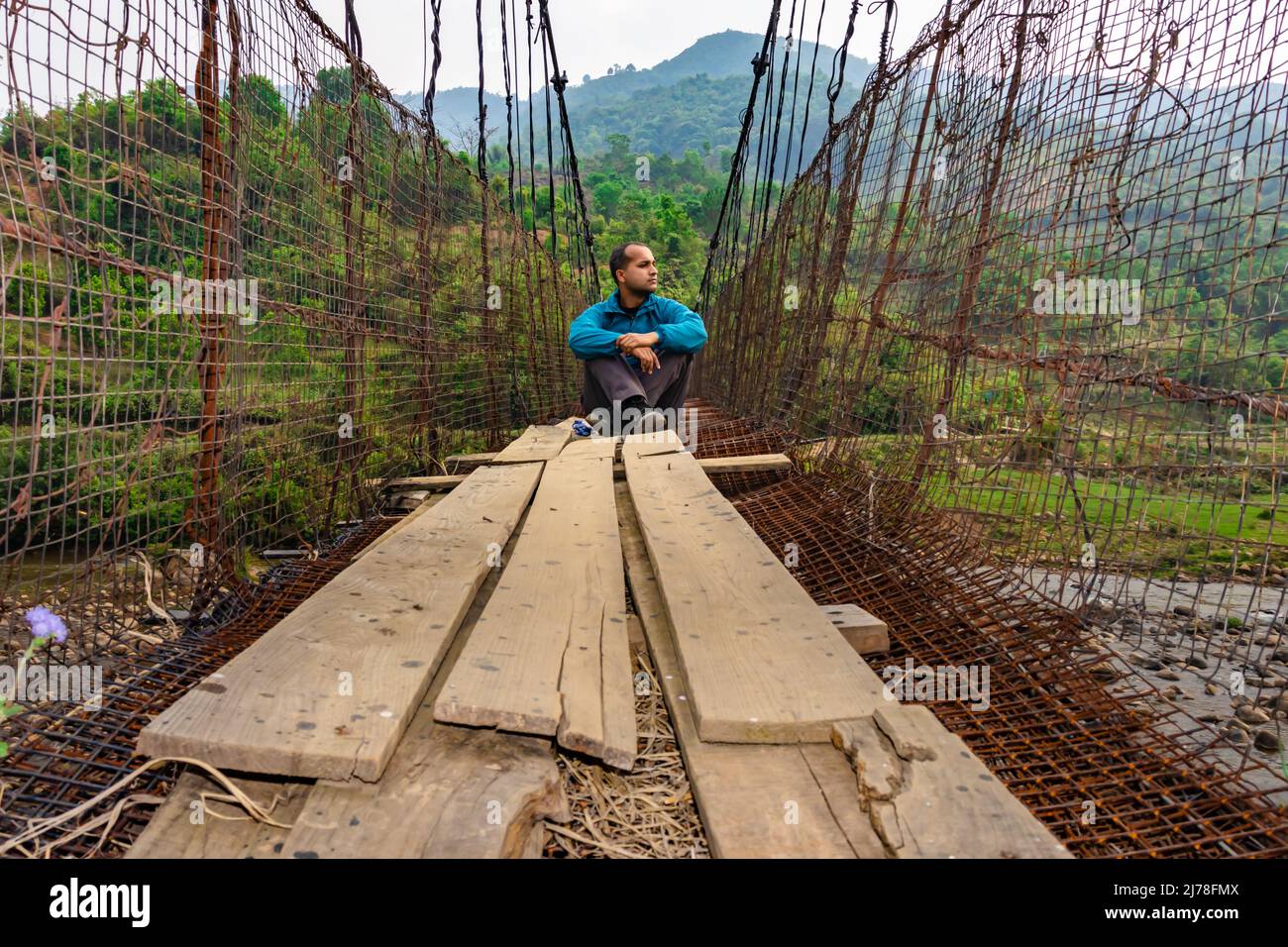 man sitting alone at vintage iron suspension bridge from low angle image is taken at nongjrong meghalaya india. Stock Photo
