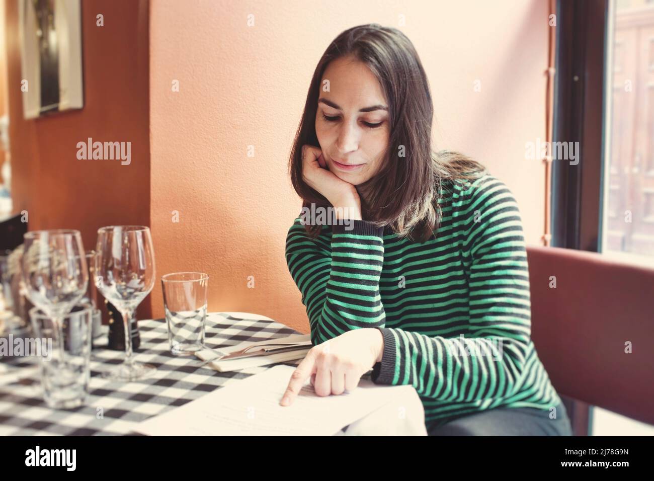 One attractive young lady sat at a table reading the menu in a bistro restaurant on her own Stock Photo