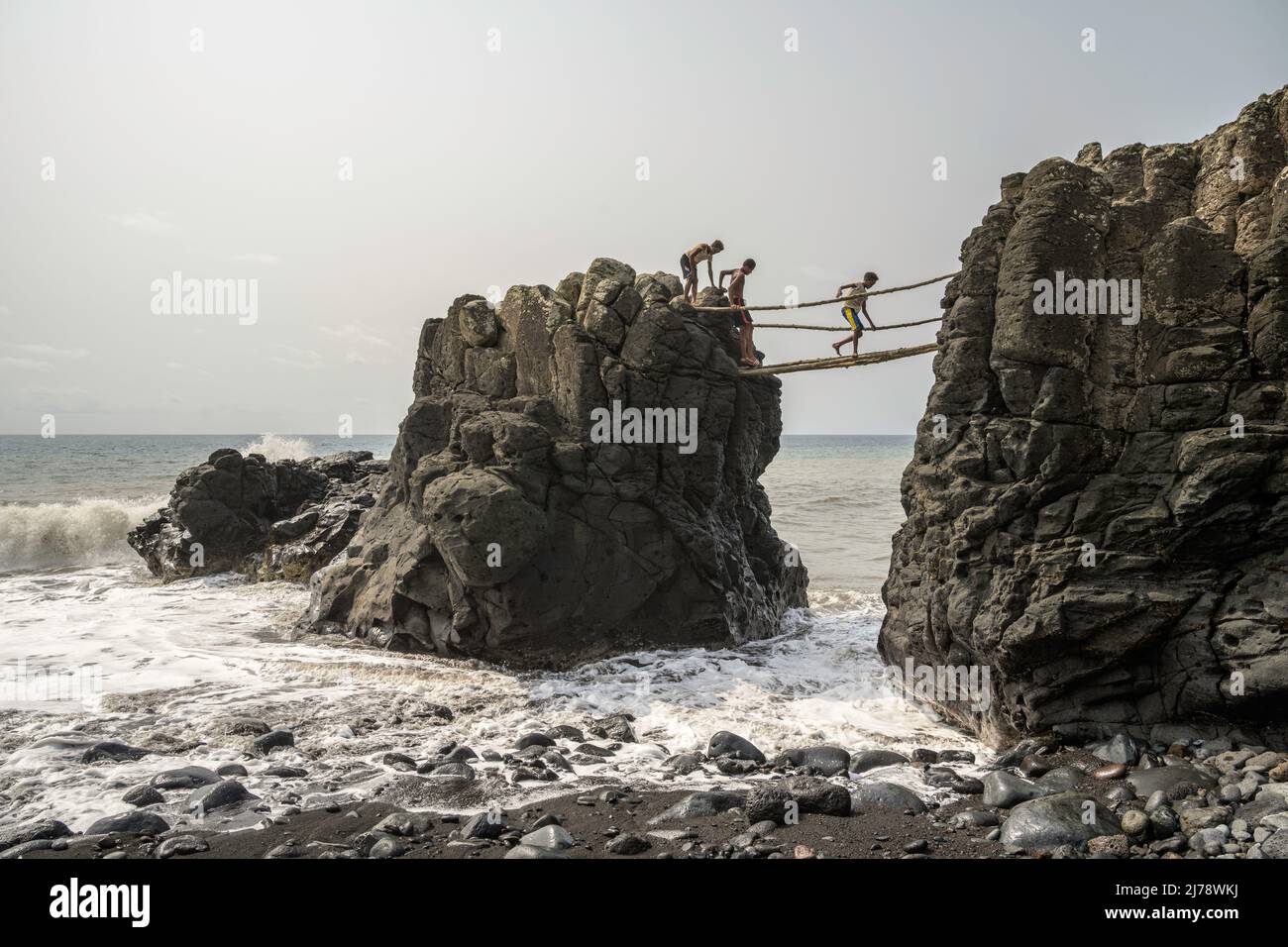Children crossing a precarious bridge made of logs while the waves beat on the rocks. Stock Photo