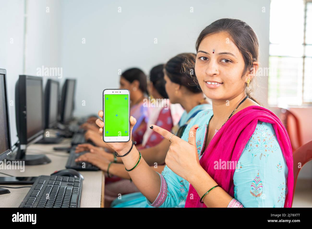 Happy women showing green screen mobile phone by pointing finger while lookingcamera at computer class - concept of online booking, advertisement and Stock Photo