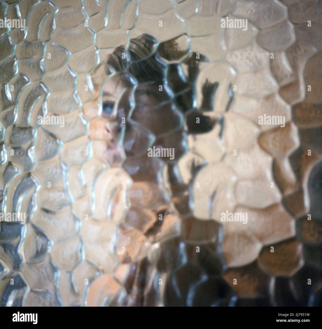 1970s, historical, the outline figure of a young woman behind a glazed, honeycomb patterned or textured glass window. This type of glass is known as figured or patterned rolled glass and was a popular style of glass used in the home in this era. Stock Photo