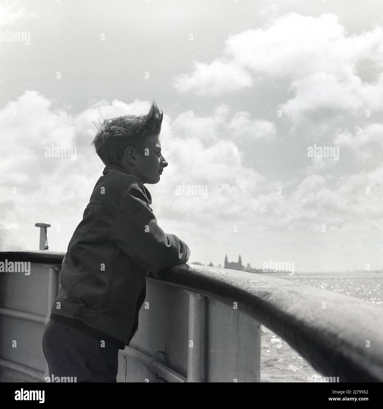 1970, historical, a boy on a boat, leaning on the wooden rail looking out, on the River Mersey, Liverpool, England, UK. The Royal Liver Building can be seen in the far distance. Stock Photo