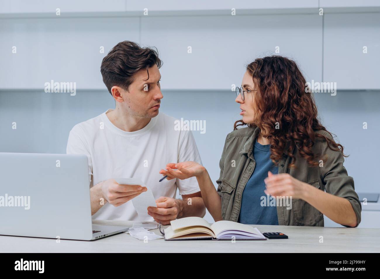 Husband and wife calculate the expense of finances sitting at home in the kitchen at the table and write it down in a notebook, a woman with checks in her hands makes excuses, a man gets upset Stock Photo