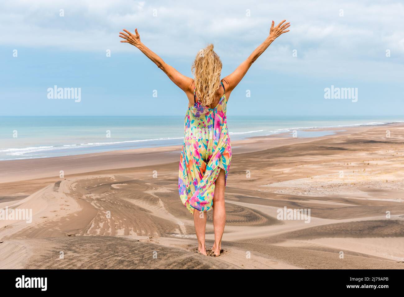 Back of a blonde woman standing on the desert gesturing freedom with the arms while facing the sea. Stock Photo
