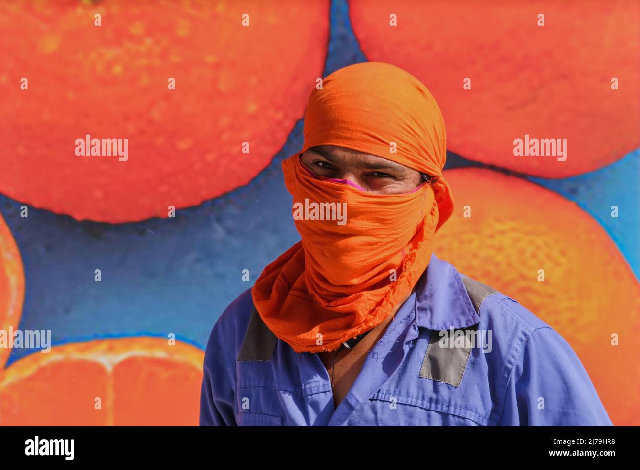 Selective focus to face of young overworked middle eastern street worker man in orange headdress saving from unbearable heat near colorful blue wall  Stock Photo