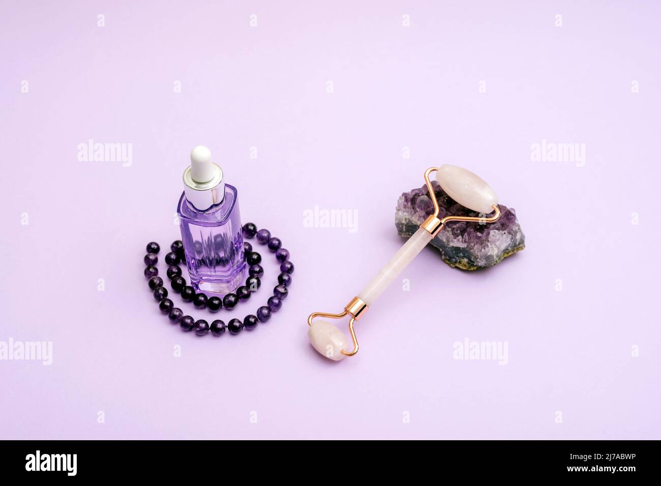 Luxury cosmetics, serum and facial roller with amethyst crystal and necklace on light purple background. Anti-aging skin care concept. Stock Photo