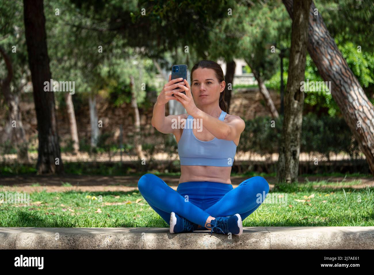 Pretty sporty woman sitting crossed legged in the park taking a selfie on her phone. Stock Photo