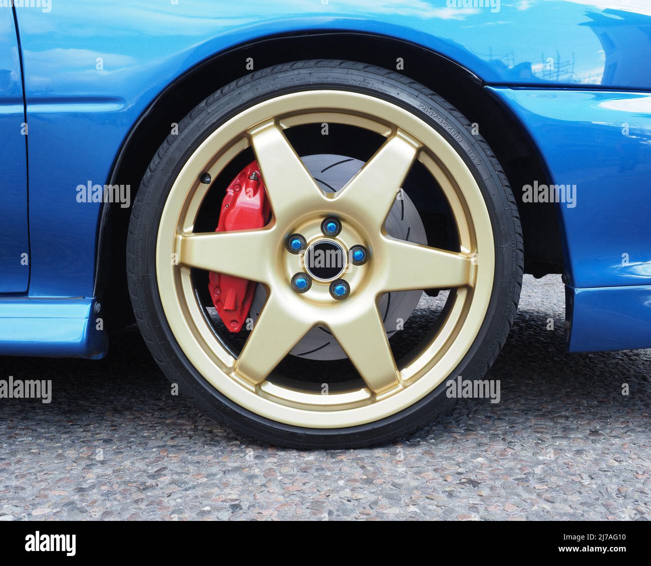 Close up of a 6 spoke metallic gold alloy wheel, 4 piston red brake calipers and grooved brake discs. Metallic blue car on tarmac road. No logos. Stock Photo