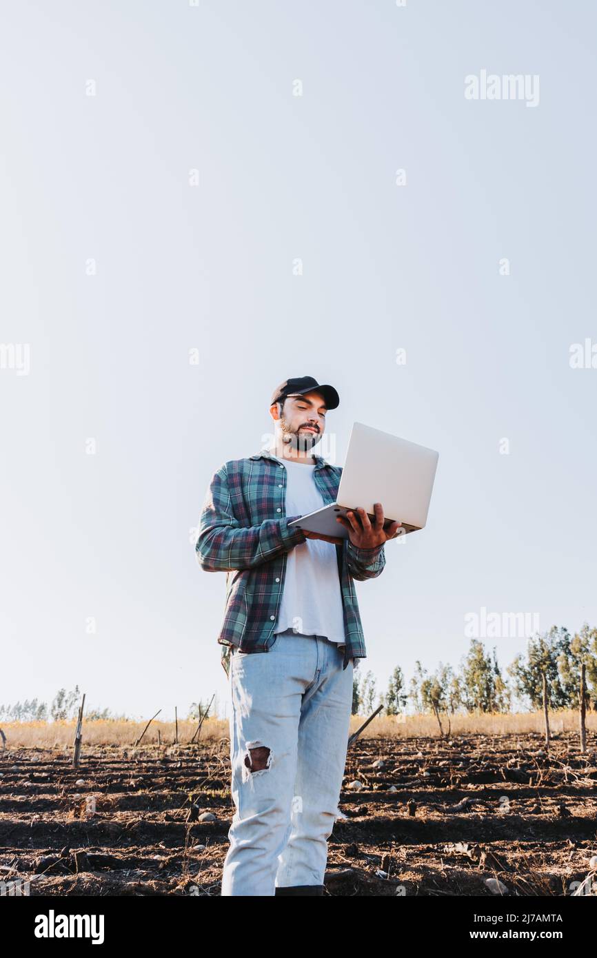 Young latin farmer man teleworking on his laptop in the middle of his farmland . Agricultural sustainability Stock Photo