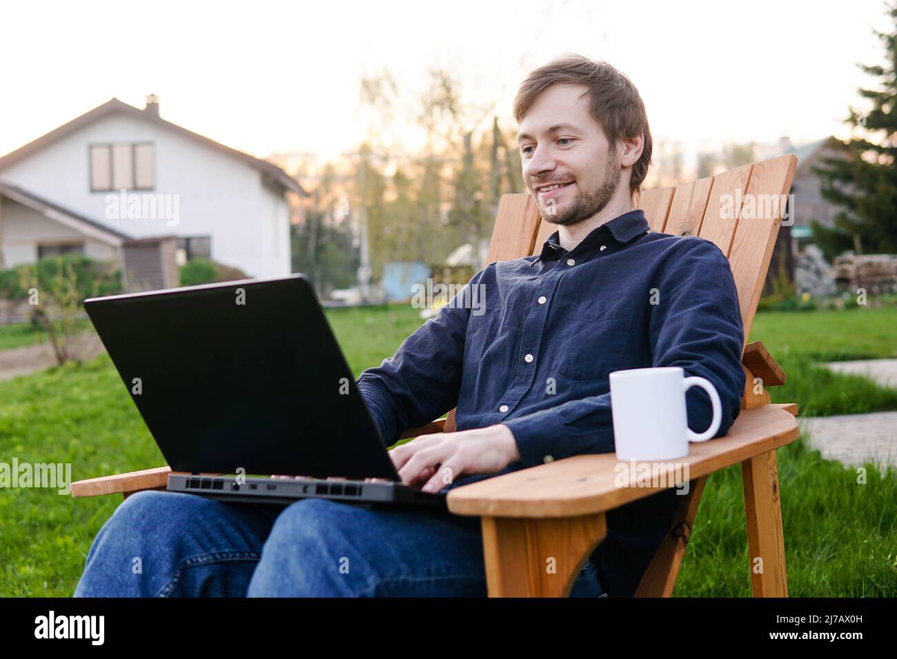 Young man freelancer sitting in wooden chair with laptop working outdoors in garden. Stock Photo