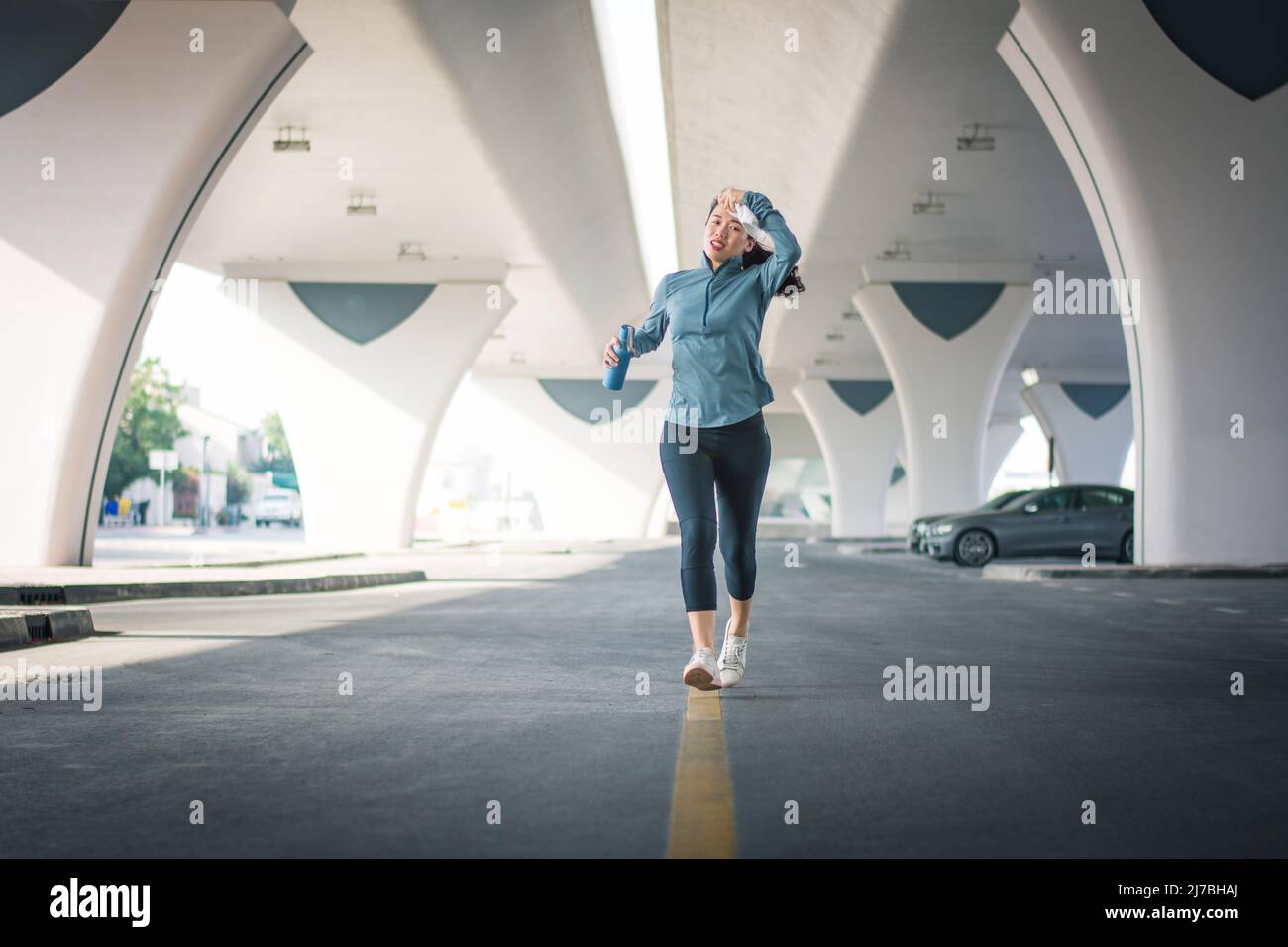 Asian woman jogging in the city at car park in urban area and wiping away sweat. Running and healthy outdoors time. Fit female person doing sport acti Stock Photo